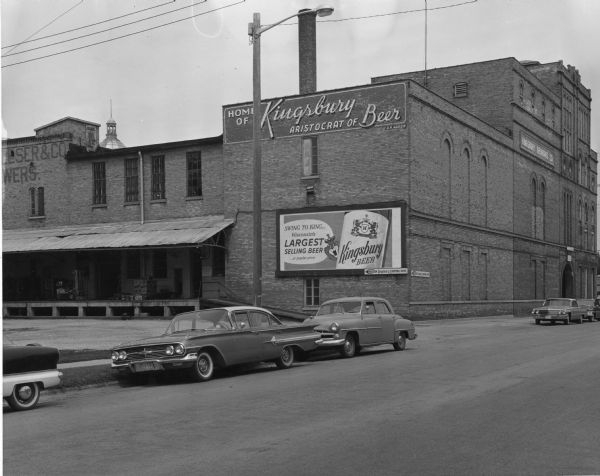 View across road towards loading dock of the Kingsbury Brewing Company. Printed on the side of the building are two advertisements for Kingsbury Beer.