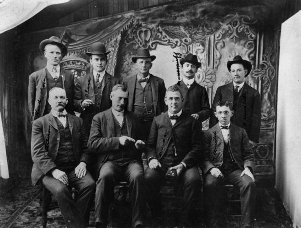 Studio portrait in front of a painted backdrop of what is probably a group of pearl buyers. The man at front left is identified as Ed Kittelson, a prominent Wisconsin pearl buyer. One man points to a small object in his hand and another holds an object with what appears to be two pearls on it.
