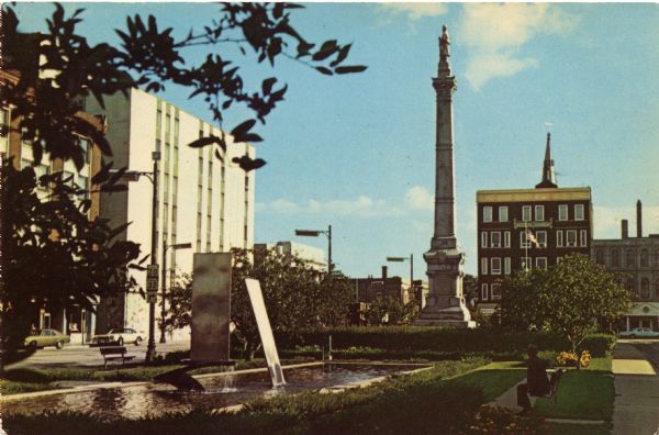 Photographic postcard view of Monument Square looking south. In the bottom right corner, a man sits on a bench looking at the fountain. The Civil War Soldiers' Memorial statue stands center right. Racine Motor Inn can be seen center left.