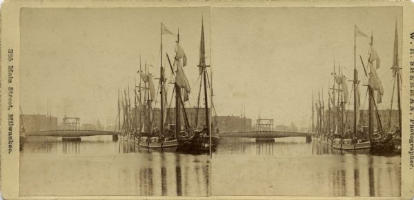 Stereograph of lumber hooker ships in the Racine harbor on the Root River. The 4th St. bridge spans the river at left and the Racine Agricultural Works building can be seen behind the ships.
