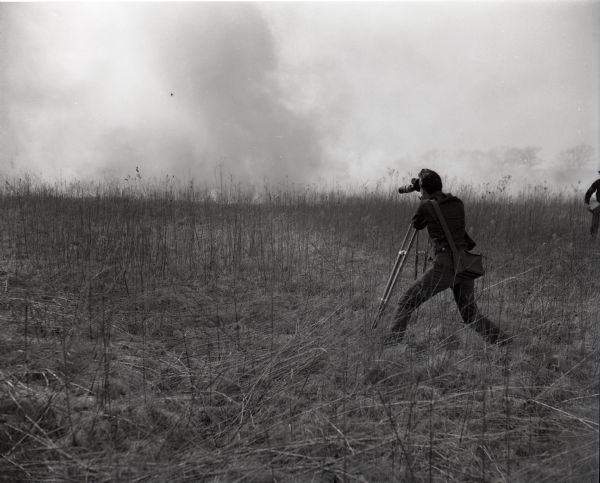 Disney film crew filming a prescribed burn at the University of Wisconsin-Madison Arboretum for inclusion in the film <i>The Vanishing Prairie</i>. A cameraman with a camera on a tripod stands among dry grasses as smoke from the burn rises in the distance.