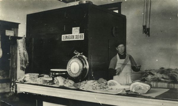 Interior view of a butcher shop, with a male butcher sharpening a knife behind a counter lined with cuts of meat, sausages and a scale. Behind the counter on a wood cabinet is a sign reading "Oleomargarine Sold Here" and a 1911 oleo tax stamp.