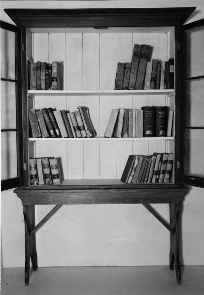 The original collection of fifty books that formed the foundation of the Wisconsin Historical Society Library in 1853. The books are displayed on shelves in a wall cabinet with the glass doors opened.
