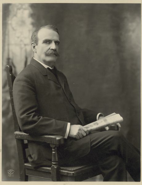 Studio portrait of Henry Clay Payne, who is seated in a chair and holding a newspaper dated August 7, 1902.