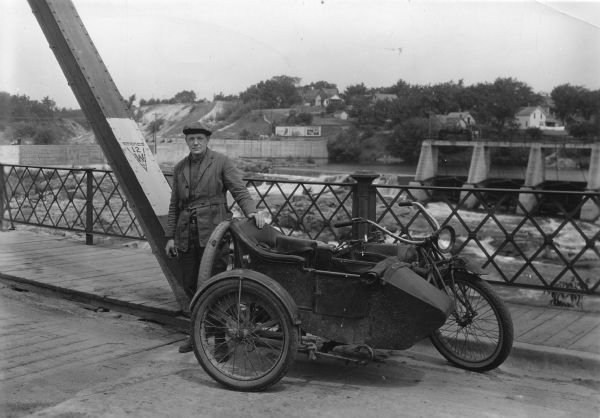 Huron Smith standing next to a motorcycle with a sidecar on a highway 12 bridge in Black River Falls. A dam across the Black River Flowage can be seen in the background.