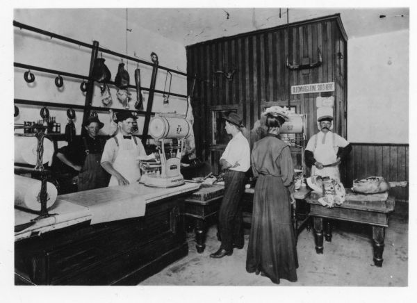 A man and a woman stand on the customer side of the counters. Phil Hof stands with a large knife behind a tabled covered with meat, while Sam and Herman Hof stand at left weighing cuts of meat. There is a sign declaring "Oleomargarine Sold Here" along with an oleo tax stamp on the wall behind the butcher.