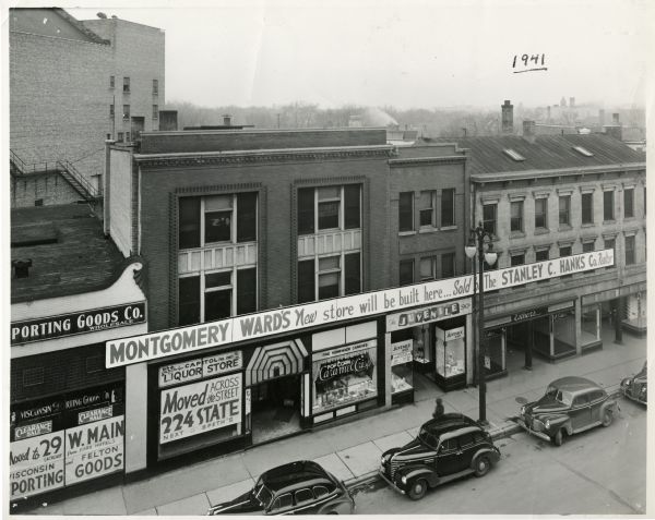 Elevated view of the south side 200 block of State Street. A large sign announces the coming of a new Montgomery Ward's store on the site. Stores shown include Wisconsin Sporting Goods Co. (moved to 29 W. Main), Capitol Liquor Store (moved to 224 State St.), Caramel Crisp candy store, The Juvenile Shop, and Esther's clothing store. There are cars parked along the curb.