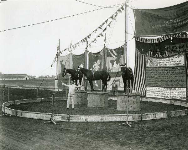 Three performing horses stand on platforms in a ring as their trainer gestures proudly in the foreground. Behind the ring are banners decorated with horses, cows, and a clown. A sign advertises the Fun on the Farm show (of which this performance was a part) and another, titled <i>Civilization at Its Worst</i> details arguments against use of oleomargarine. Another man stands in the background outside the ring.