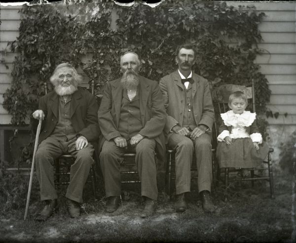 Informal outdoor group portrait of, left to right: William (holding cane), August, Alex and Edgar Krueger sitting in chairs in front of a trellis on the side of a house.