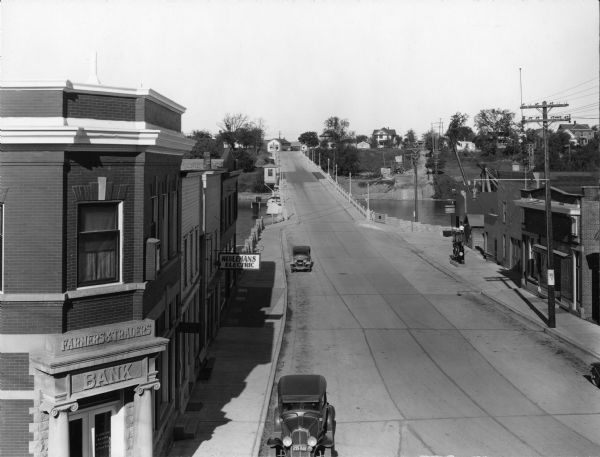 Elevated view of High Street (Highway 96) and  the bridge over the Fox River at Wrightstown. Farmers & Traders Bank is at left, a sign for Meuleman's Electric can be seen a couple doors down. Two automobiles are parked along the curb on the left side of the street. A Chevrolet sales and service station with curbside gas pumps is on the opposite side of the street. A residential area can be seen across the bridge.