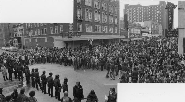 Composite photograph of an elevated view of an anti-Vietnam War protest at the intersection of Lake and State Streets looking east up State Street. A large group of protestors is gathered on State Street and a line of police officers in riot gear block entry to the University of Wisconsin campus. Businesses visible in the photograph include Brown's Books, Rennebohm's, Warner Medlin, Antoine's 5th Avenue, Discount Records and Taco Grande.