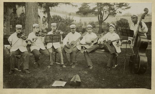 String band consisting of seven inmates at Waupun State Prison. Three seated men hold guitars, three others hold violins and one standing man holds a cello. All wear prison uniforms. They are posed in the grass near a tree in the prison yard.