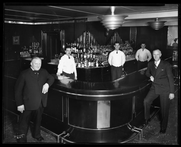 Interior photograph taken from the corner of a large black bar. Two men wearing suits are leaning against the bar, and three bartenders are standing behind it. There is a large display of liquor bottles and glassware, and a cash register is behind the bar.