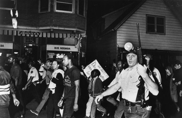 Father James Groppi (center) and the NAACP participate in a civil rights march. Police are armed and escorting the protestors. The marchers are passing an establishment named Johnny's.