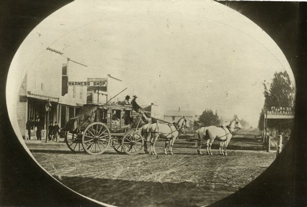 Photographic postcard of a stagecoach pulled by four horses in the road in front of a harness shop. Two men sit in the upper seat. A handful of people stand on the sidewalk at left. A shop selling apples, flour and feed is at right.