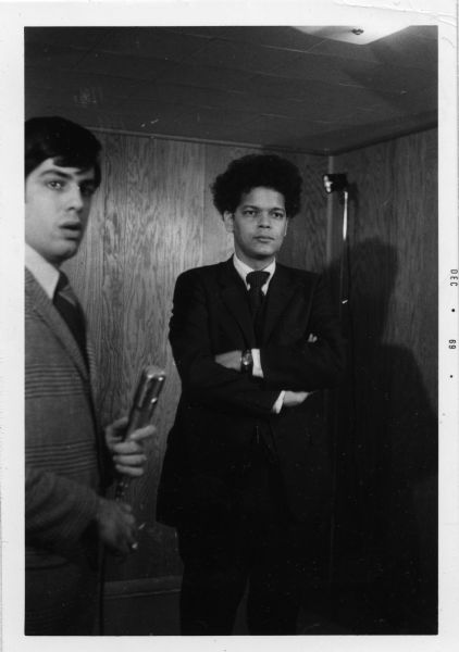 Julian Bond wearing a suit and standing in a wood paneled room with his arms crossed. Another man is standing at left holding a microphone.