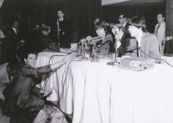 Radio disc jockey "Beatle" Bob Barry squats in front of the table where The Beatles are seated for a press conference before a concert.