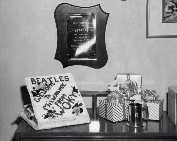 A table and wall display a cake, presents, and a plaque honoring the arrival of The Beatles. Icing on the cake reads "Beatles Welcome To Milwaukee From WOKY." The plaque reads "In appreciation to The Beatles for their visit to Milwaukee September 4, 1964 presented by Milwaukee Welcomes The Beatles Club."