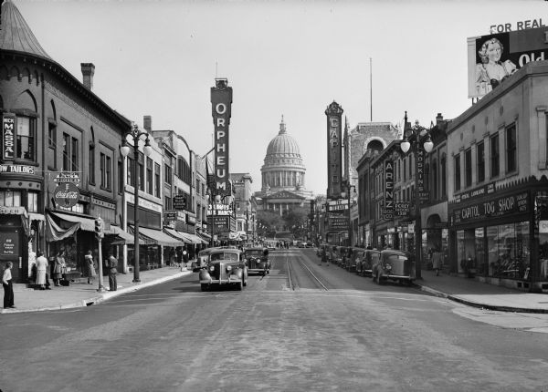 A view from the 200 block of State Street looking toward the Wisconsin State Capitol. This urban scene includes marquees for the Orpheum Theatre and the Capitol Theatre, storefronts, traffic, and pedestrians. 