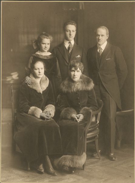 Full-length formal studio portrait in front of a painted backdrop of the family of Harold McCormick and Edith Rockefeller McCormick. From left to right, standing, are: Mathilde McCormick, Fowler McCormick and Harold McCormick. Seated from left to right are: Muriel McCormick and Edith, both wearing long coats.