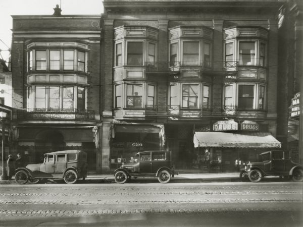 View across street of the 200 block of State Street, showing pedestrians in conversation, parked automobiles, streetcar tracks, and the storefronts of a shoe store, the Photoart House, and a Rennebohm Drug Store. This Rennebohm's store, which was variously numbered as 204 and 208 State Street, was opened in 1924 as Rennebohm's second store. At the time it was known as the Central Drug Store. The building in which it was located was built in 1909 and designed by Madison architect Ferdinand Kronenberg. Except for the store windows, the original facade is largely intact.