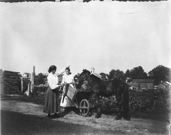 Lena and Alma Herling at their farm with a horse and a wheelbarrel. In the far background are farm buildings and what appears to be a church.