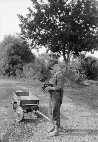 A young barefoot boy wearing striped coveralls stands on a dirt road while holding a box camera. A coaster wagon is next to him.