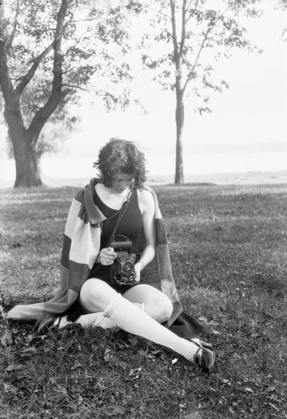 A female model poses with a camera in her lap while sitting in the grass. She is wearing a black bathing suit, a striped cape, and shoes with knee-high stockings.
