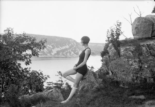 A female model poses in profile while leaning back against rocks. She is wearing a black bathing suit and knee-high stockings. In the background is a lake with a hill or bluff on the far shoreline.