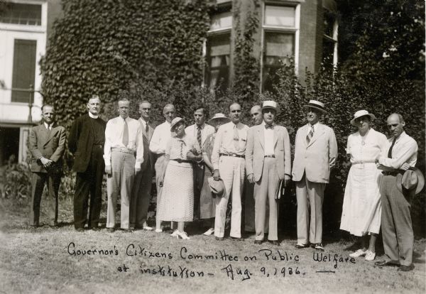 Outdoor group portrait of fourteen people who make up the Governor's Citizens Committee on Public Welfare. They are posed front of a building at the Northern Wisconsin Center for the Developmentally Disabled.