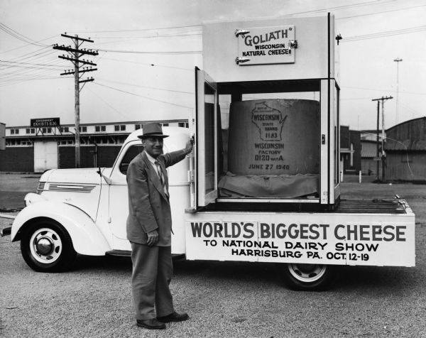 Man posed next to a truck hauling the world's biggest cheese, named "Goliath" by The Wisconsin Department of Agriculture. Probably taken at the Wisconsin State Fair Grounds prior to the shipment of the cheese to the National Dairy Show in Harrisburg, Pennsylvania and the World's Fair in New York.