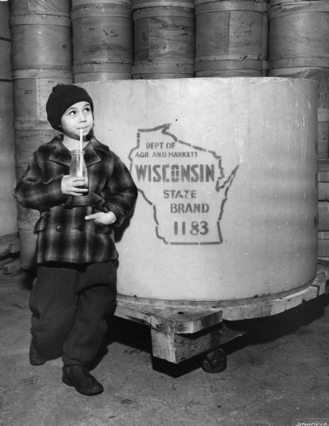 Boy dressed in coat and hat and sipping milk from a bottle poses with "Goliath," the world's biggest cheese, which is resting on a wheeled platform. Round cheese boxes are stacked in the background.