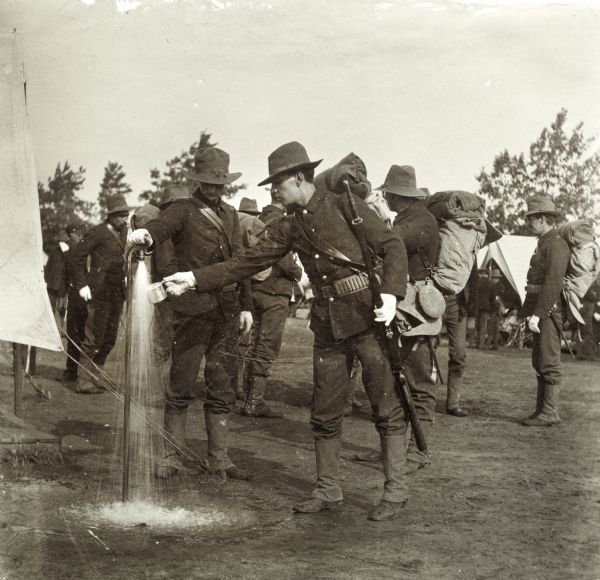 Wisconsin National Guardsmen in full gear. One man in the foreground fills a tin cup from a spigot.