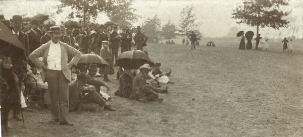 A group of visitors to Camp Douglas are gathered at the edge of an open field, perhaps observing military exercises. Several carry umbrellas.