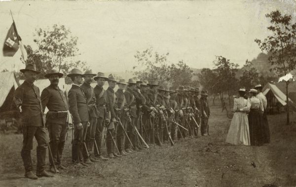 Members of the Wisconsin National Guard Light Horse Squadron, Troop A standing in a row. Three women and one man, visitors to Camp Douglas, stand facing the line.
