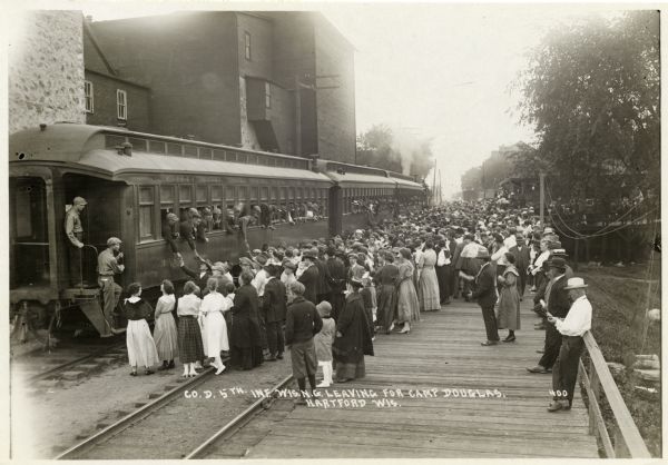Large crowd of people gathered at the train station to see off Company D of the 5th Wisconsin National Guard Infantry. The soldiers, some leaning out of the windows of the train to shake hands, were leaving Hartford for Camp Douglas.