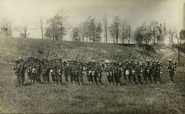 Members of the Wisconsin Light Guard standing in formation in a field. There is a hill in the background lined with trees.