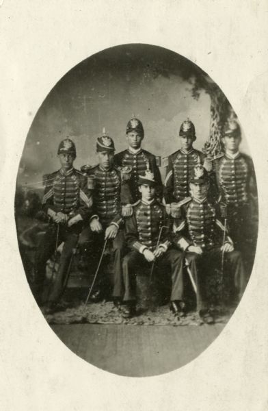 Oval group portrait of the Racine Light Guards posed in front of a painted background. Standing left to right: Arthur Mack, U.S. Grant (Corporal), Matthias Maus (?); and seated left to right: Charles Madison (Sergeant), George Graves (Captain), John Vaughan (1st Lieutenant from Milwaukee), Michael Quinn (Corporal) posed in military dress uniform.
