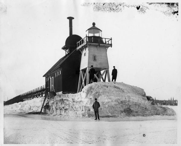 Three men pose standing on ice around North Pier lighthouse. In the background is the town along the shoreline.