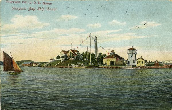 Hand-colored postcard view of the Sturgeon Bay ship canal from the bay toward shore. A sailboat is on the left and a lighthouse is on shore on the right. Caption reads: "Sturgeon Bay Ship Canal."