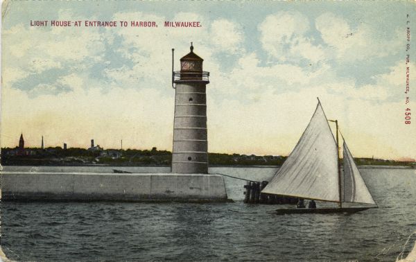 Hand-colored postcard view of a pier and lighthouse at the entrance to Milwaukee's harbor. Two people sail in a small boat to the right of the lighthouse, and the city is along the shoreline in the background. Caption reads: "Light House at Entrance to Harbor, Milwaukee."