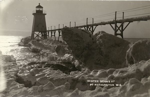 View of ice coated pier and lighthouse along the shore of Lake Michigan. Caption reads: "Winter Scene at Port Washington, Wis."