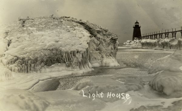 Winter postcard view of the pier and lighthouse at Port Washington seen from the icy shore of Lake Michigan. Caption reads: "Light House."