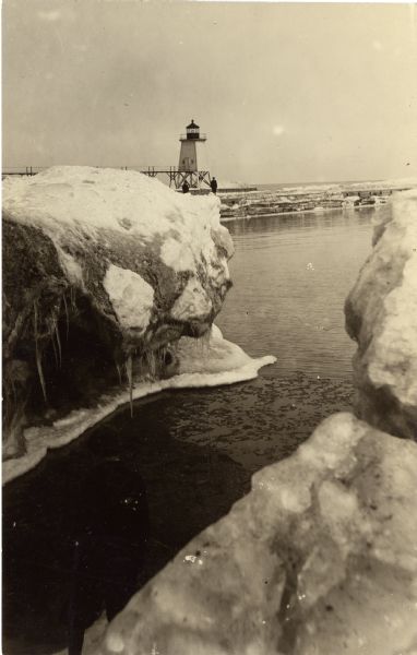 Winter view of the lighthouse at Port Washington from the icy shore of Lake Michigan. Two people can be seen standing on a pier in the distance.