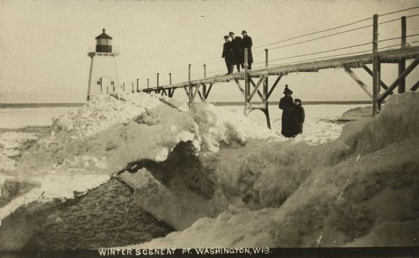 Postcard view of three men on an elevated walk on an icy pier near the shoreline. A women and young boy stand on the pier below the walk. There is a lighthouse at the end of the pier. Caption reads: "Winter Scene at Pt. Washington, Wis."