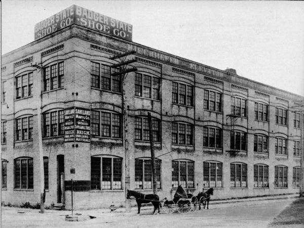 Exterior view of the building at the corner of N. 12th St. and W. St. Paul Avenue that housed Badger State Shoe Co. and Saveland Manufacturing Co. Two horses are hitched to empty carriages standing in the road in front of the building.