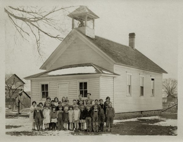 Students and their teacher, Gladys Hofseth (back row, far right), pose outside Eagle Corners School. The school is a wood frame building with an enclosed porch and a small bell tower on the roof. There is a trace of snow on the ground and trees are bare. A barn and a water pump can be seen in the background at left.