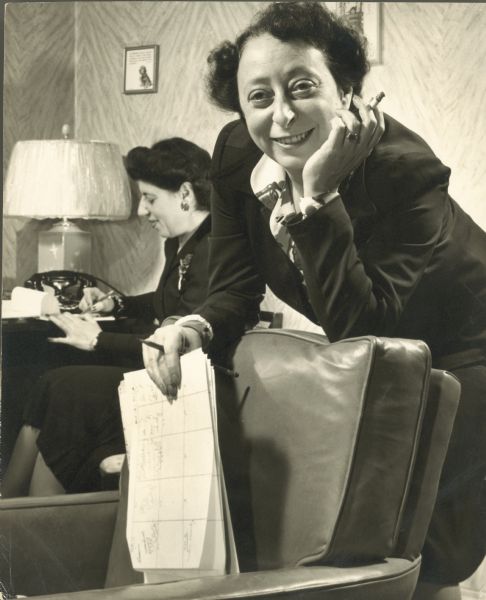 Irna Phillips, with her friend and secretary Rose Cooperman. Irna is holding papers and smoking a cigarette. Rose is sitting in the background writing.