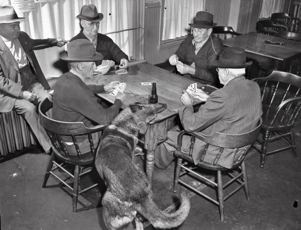 Four men playing cards at a table with a fifth man seated on a radiator observing at left. In the foreground, a dog clears an empty beer bottle from the table. The dog, Fido, was trained by the bar owner, Ernie Thierstein, to remove beer bottles from tables when and only when they are empty. The men at the table are identified as (left to right): John Marty, Henry B. Hefty, Casper Zentner, and J. Henry Zentner. The man on the radiator is unidentified, possibly Ernie Thierstein.