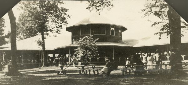 View across lawn towards a large shelter at Bernard's Park filled with people. Other people relax at picnic tables in under trees in the foreground.